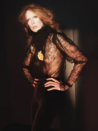 Model Raquel Zimmermann posing with hands on hips wearing a sheer black lace blouse with a gold brooch against a dark background.