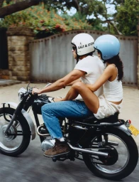 Man and woman wearing helmets riding a vintage motorcycle on an outdoor paved road.