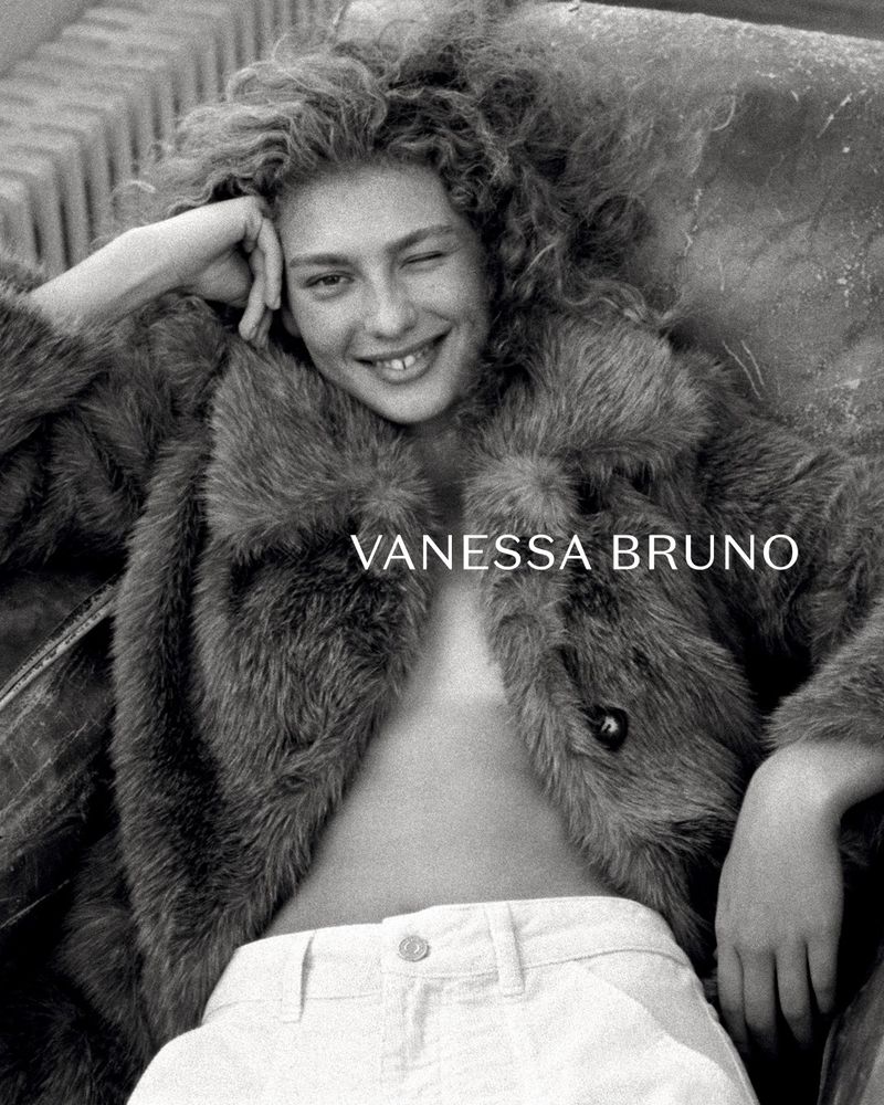 Model Dorit Revelis winking while wearing an open plush fur coat and white trousers in a black and white portrait.