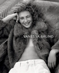 Model Dorit Revelis winking while wearing an open plush fur coat and white trousers in a black and white portrait.