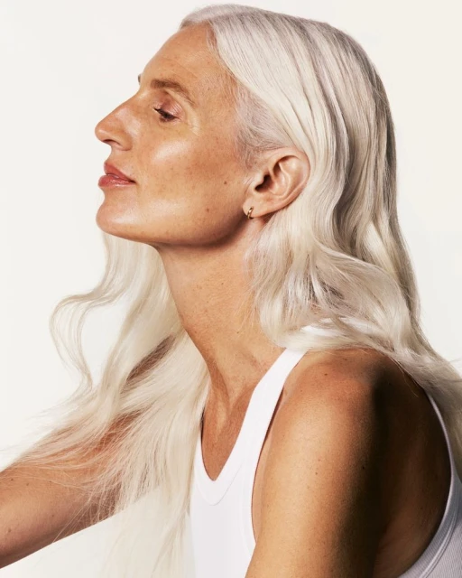 Profile portrait of a mature adult woman with flowing long white hair wearing a white tank top in a light studio setting.
