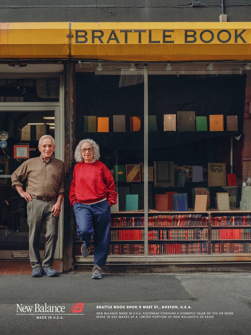 An older man and woman pose outside the yellow awning and book display of Brattle Book Shop wearing New Balance sneakers.