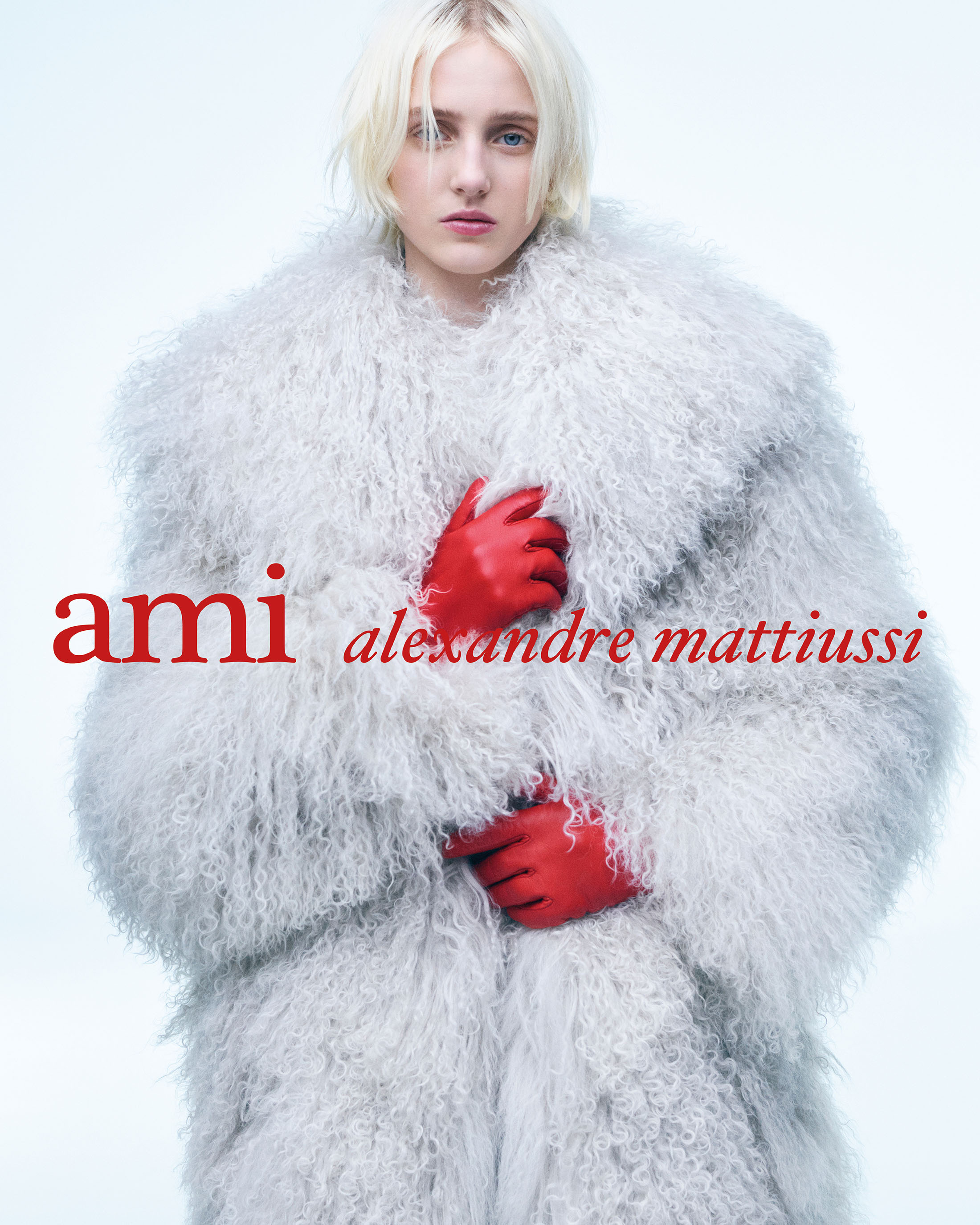 Model wearing a voluminous light grey shearling coat and vibrant red leather gloves against a bright seamless white background.