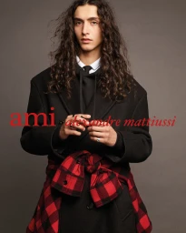 Model with long curly hair wearing a black oversized coat with a red buffalo plaid shirt tied at the waist in a studio setting.