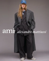 Model wearing an oversized grey overcoat, dark double-breasted suit, and blue baseball cap against a grey studio backdrop.
