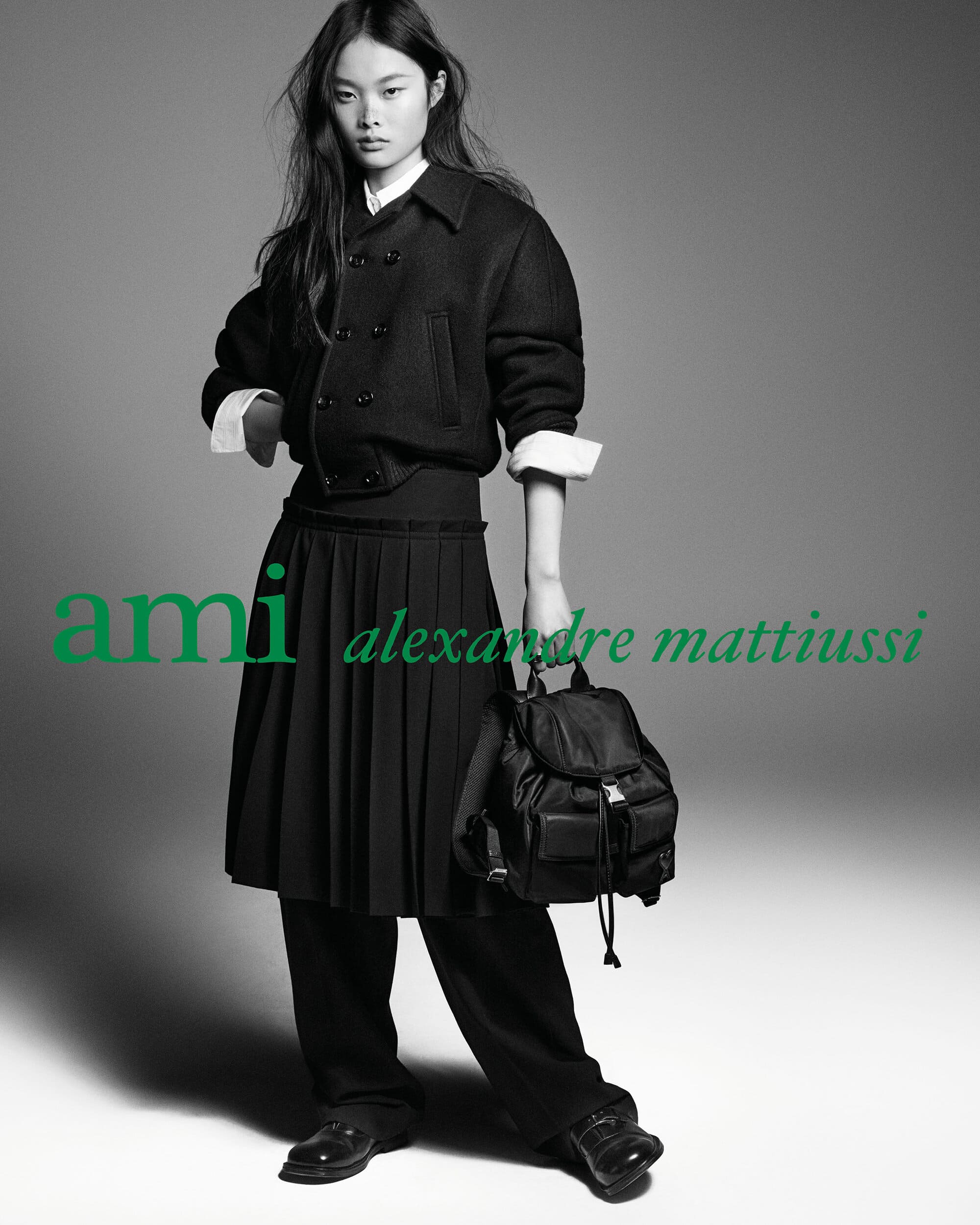 Jingwen Jia wearing a black jacket and pleated skirt holding a black backpack in Ami Paris Fall 2026 campaign.