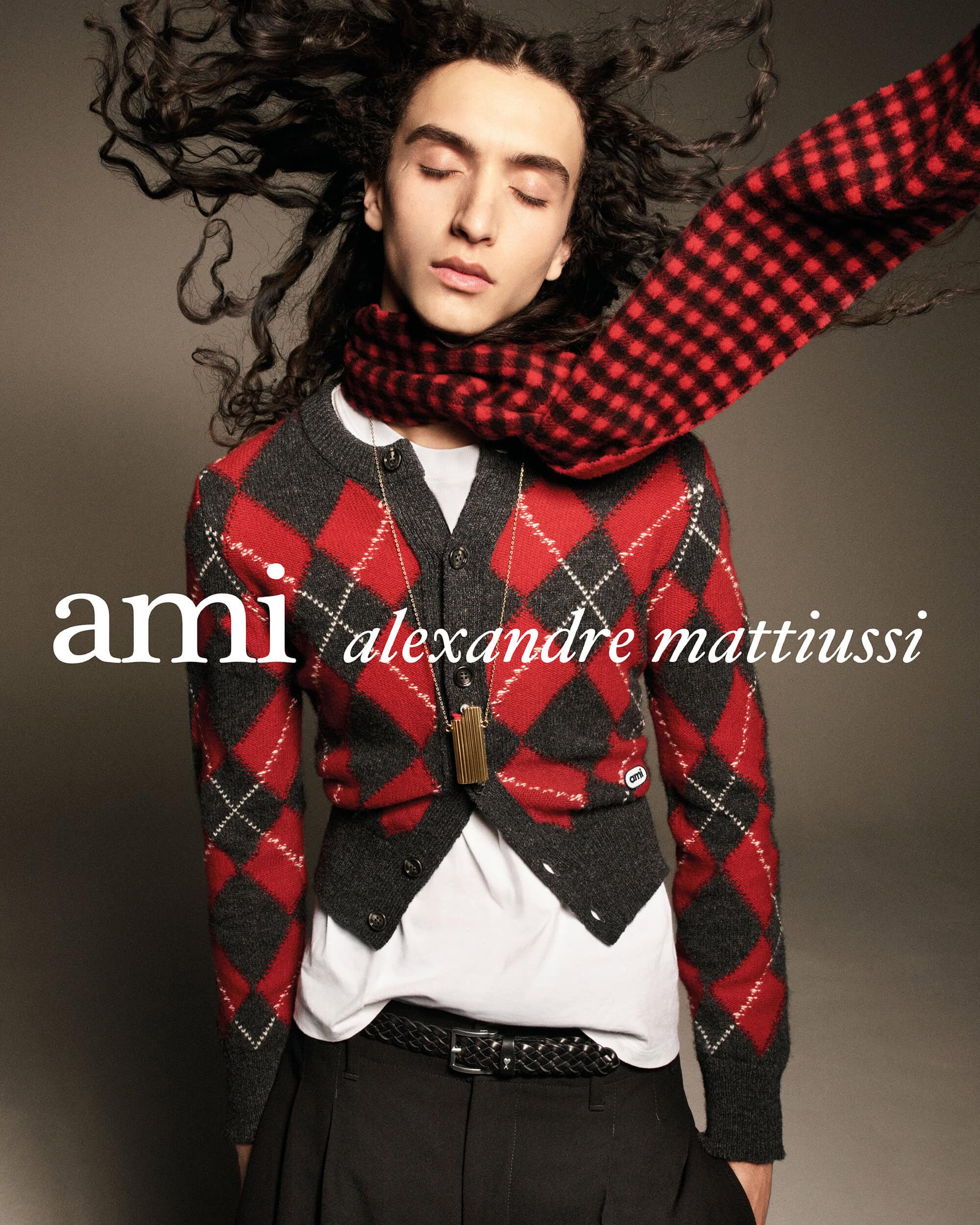 Model with windblown hair wearing a red and grey argyle knit cardigan and checkered scarf against a neutral studio backdrop.