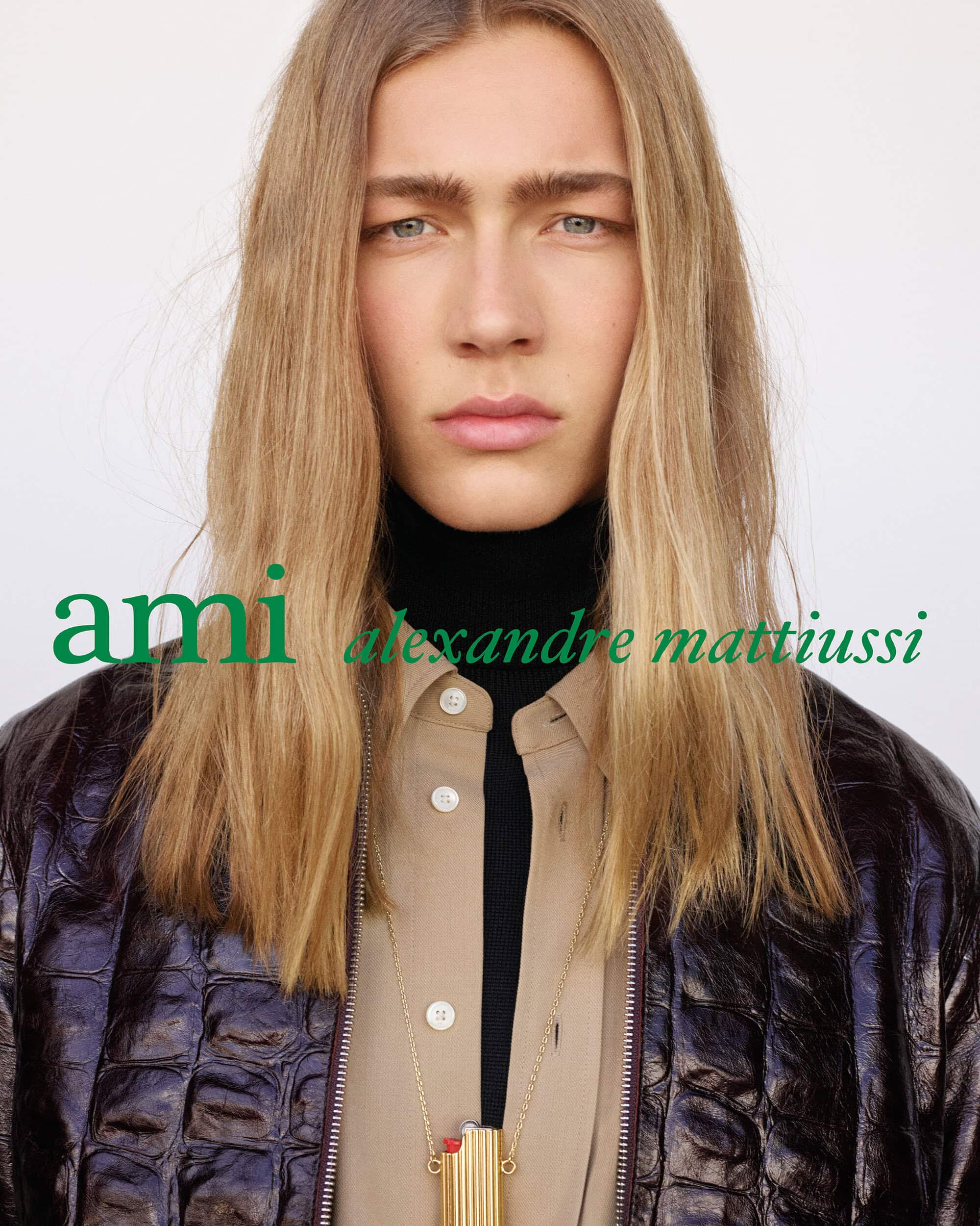 Model with long hair wearing an embossed leather jacket, beige shirt, and lighter necklace in front of a white backdrop.