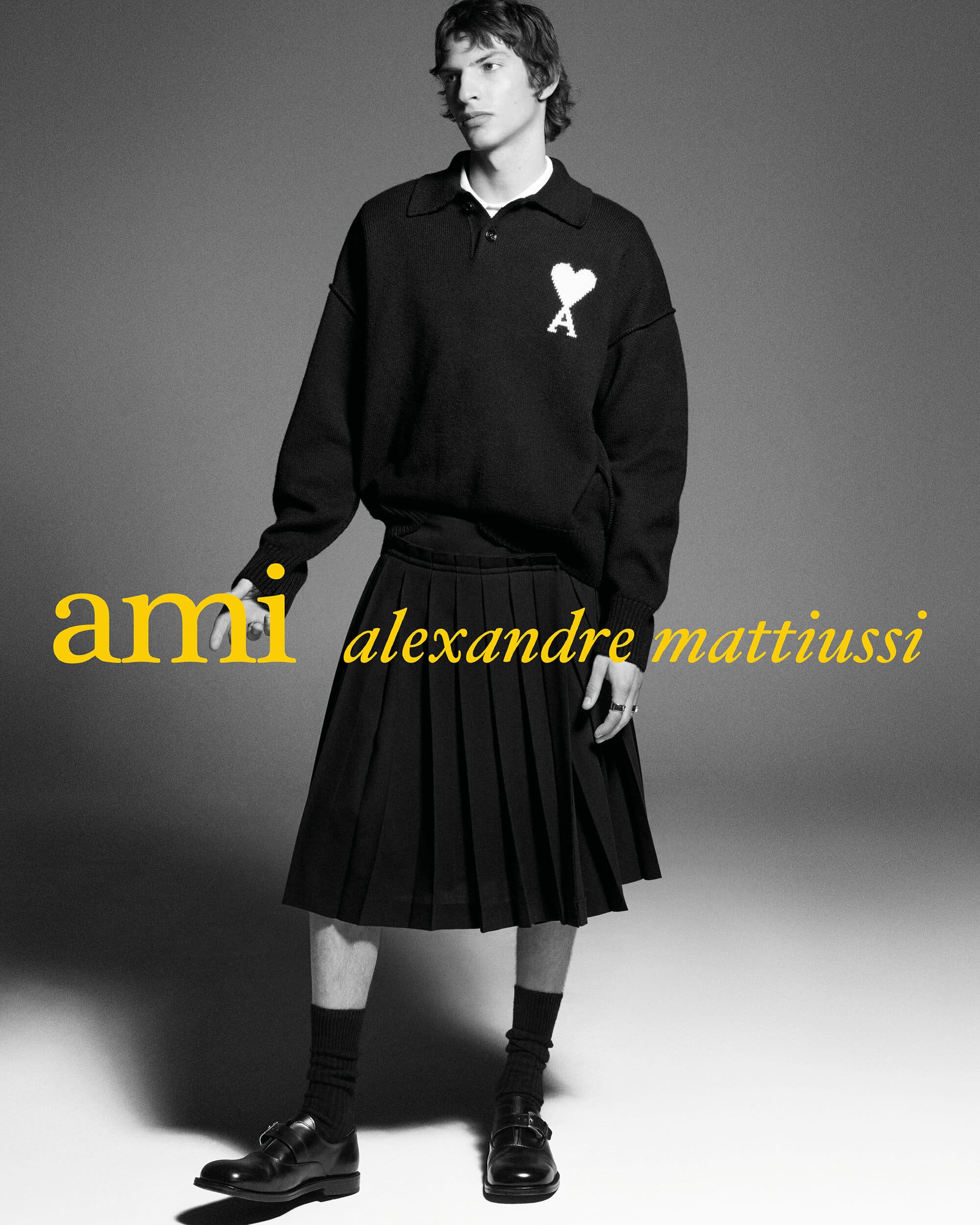 Male model wearing a black knit polo sweater with a white heart logo, a black pleated skirt, and black dress shoes.