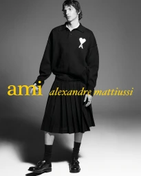 Male model wearing a black knit polo sweater with a white heart logo, a black pleated skirt, and black dress shoes.