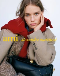 Model Rebecca Leigh Longendyke in a beige wool coat, red knit layer, and black leather handbag against a white studio background.