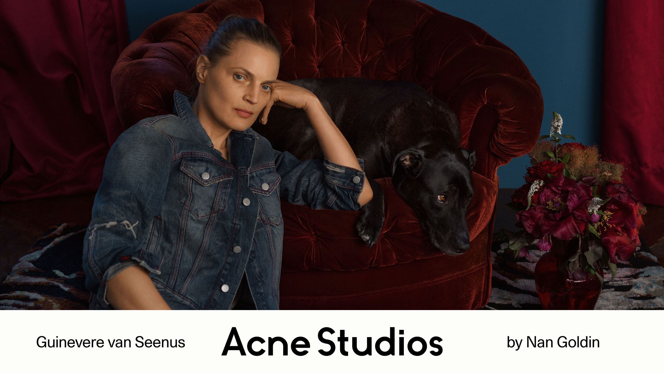 Guinevere Van Seenus in a denim jacket leaning against a red velvet armchair with a black dog.