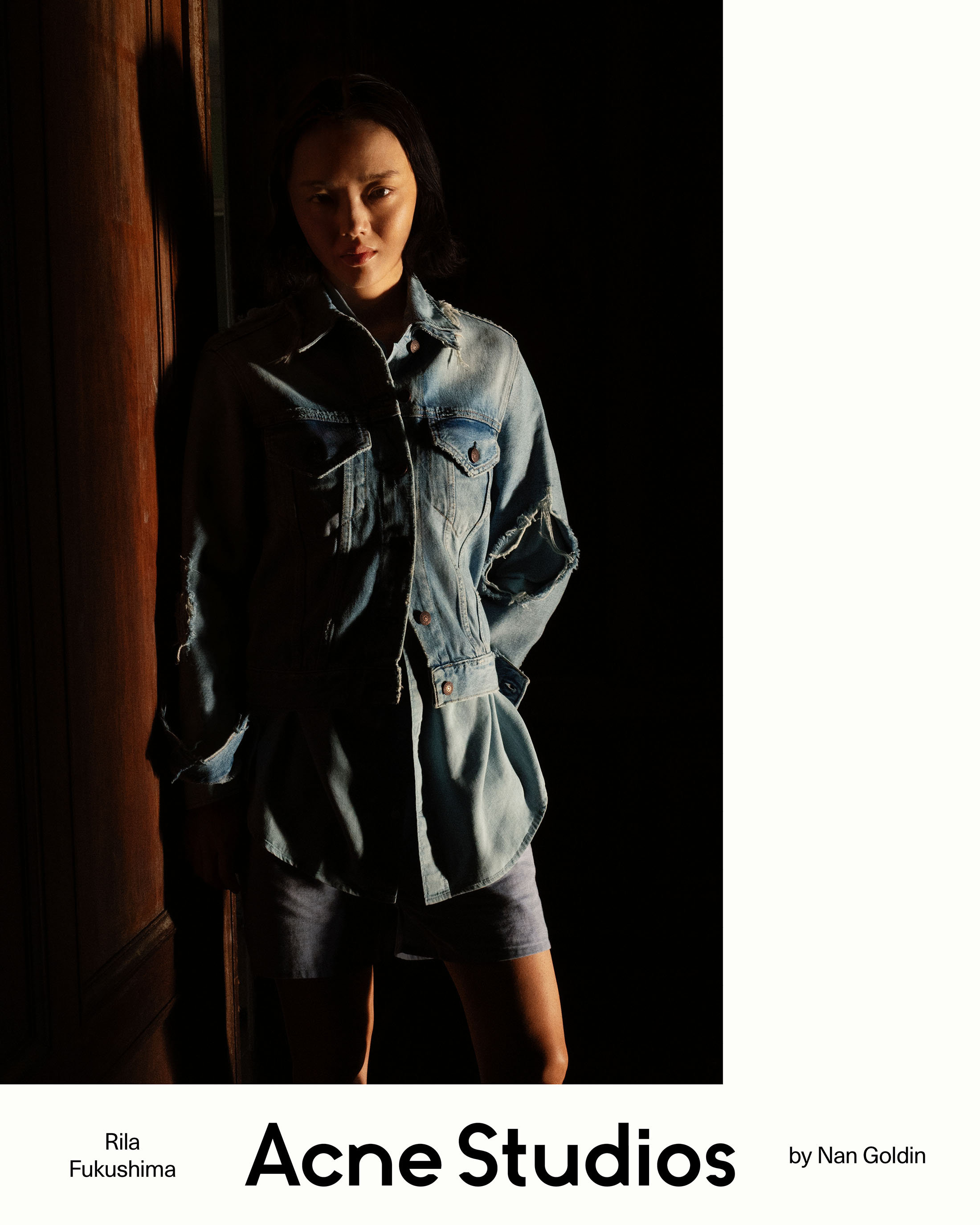 Rila Fukushima posing in a distressed light-wash denim jacket and shorts under dramatic chiaroscuro lighting for Acne Studios.