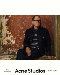 Peter Schlesinger seated in a dark denim jacket and glasses against a distressed patterned wall for Acne Studios.