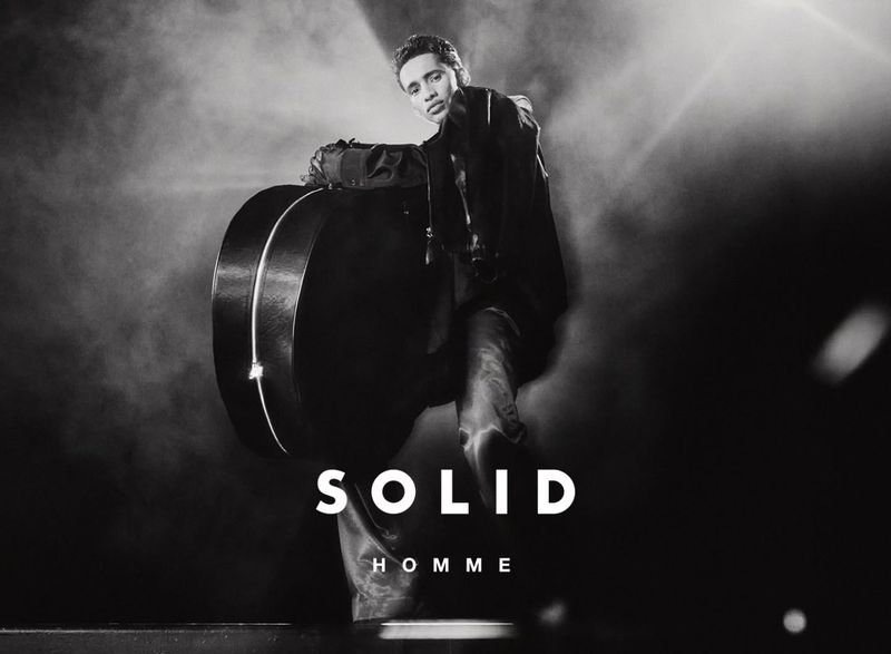 Black and white portrait of a young adult male with a guitar case, dressed in a draped jacket, against a dark, smoky background.