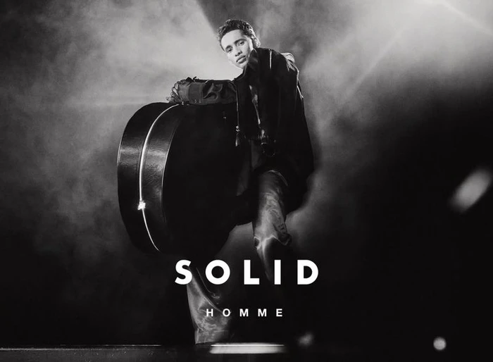 Model Abdallah El Farjani poses with a large rounded hard case under dramatic spotlight for the Solid Homme F/W 2025 campaign.