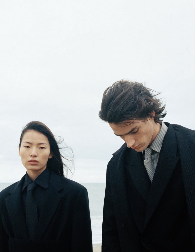 Linna Shi and Noah Herbst wearing dark tailored coats and ties against a pale overcast sky for ICICLE.