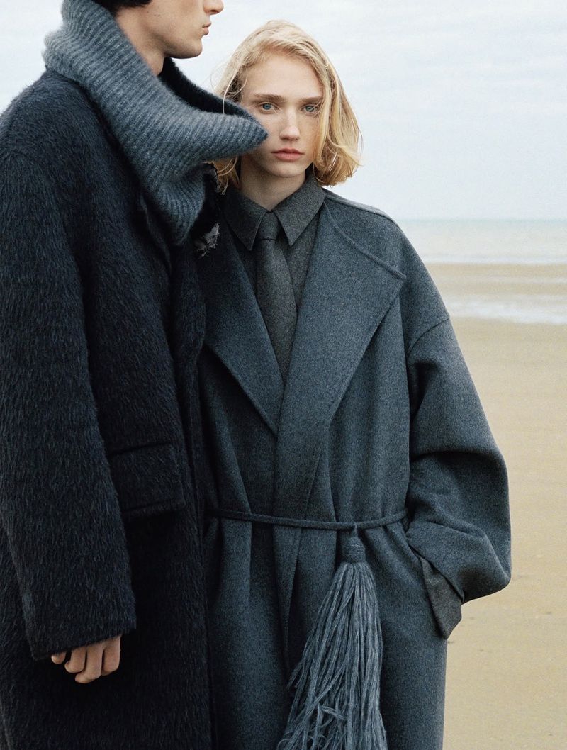 Models standing on a sandy beach wearing oversized dark wool coats, knitwear, and winter tailoring.