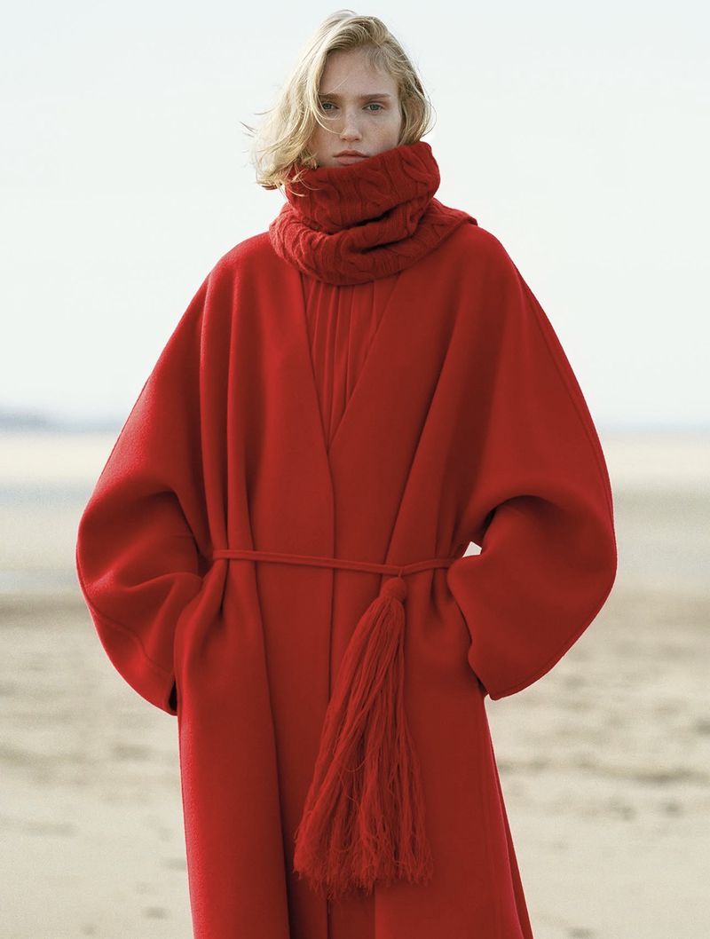 Model wearing a voluminous red wool coat with a matching knit scarf and tassel belt on a sandy beach.