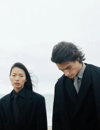 Linna Shi and Noah Herbst wearing dark tailored coats and ties against a pale overcast sky for ICICLE.