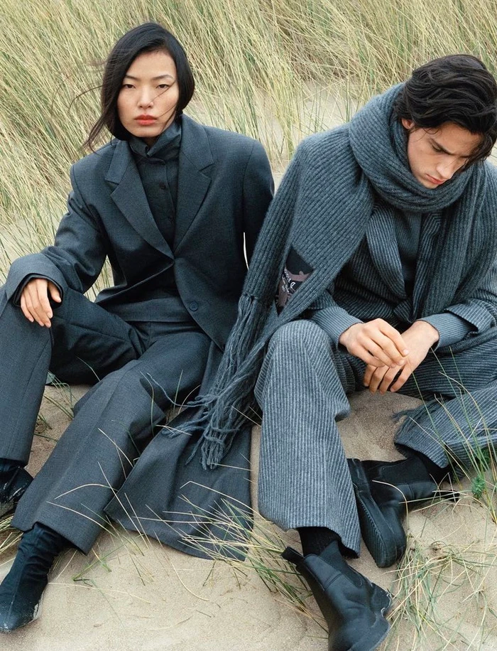 Two models wearing layered grey winter apparel seated outdoors on sandy dunes with tall grass
