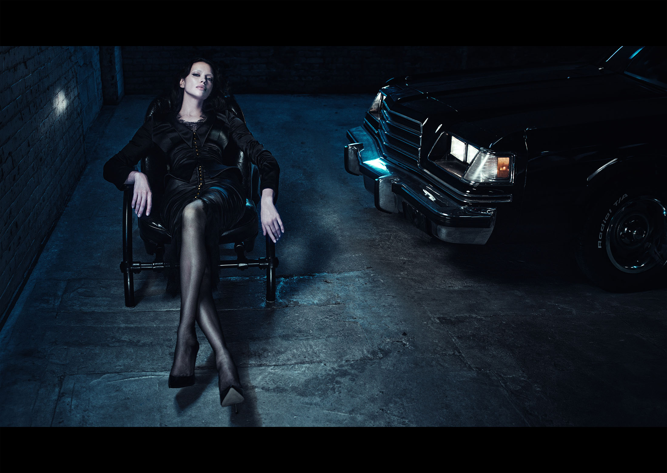 Irina Shayk seated in a black armchair wearing a dark dress next to a vintage black car in a dimly lit garage.