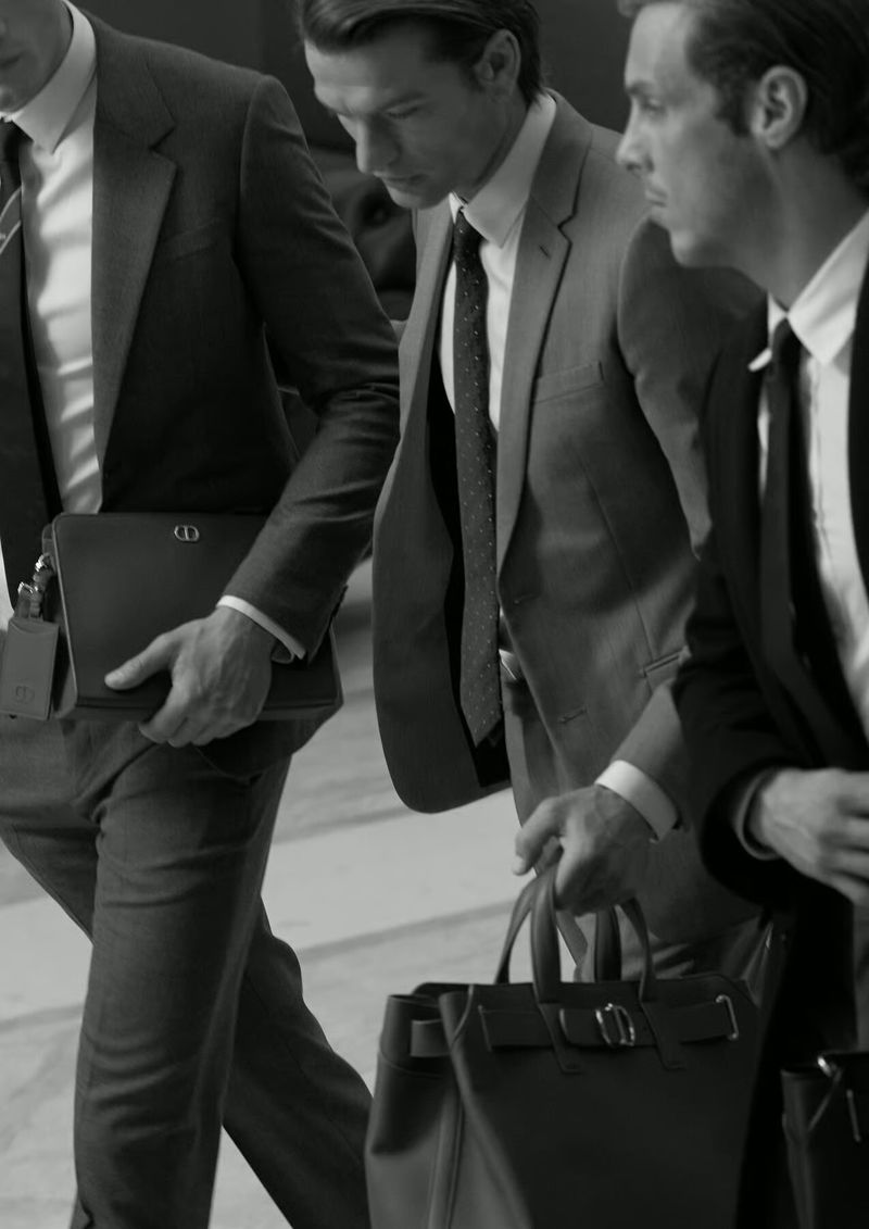 Three men in tailored suits walking side-by-side, carrying leather briefcases and tote bags. Shot in black and white.