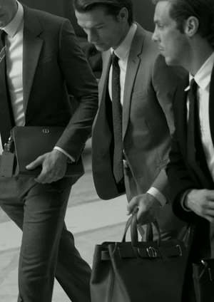 Three men in sharp tailored suits and ties walk in motion while carrying leather bags in a black and white shot.