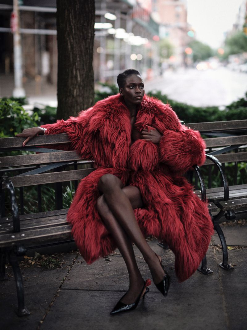 Model Ajah Angau Jok in a voluminous red shaggy fur coat and black heels seated on a wooden park bench.
