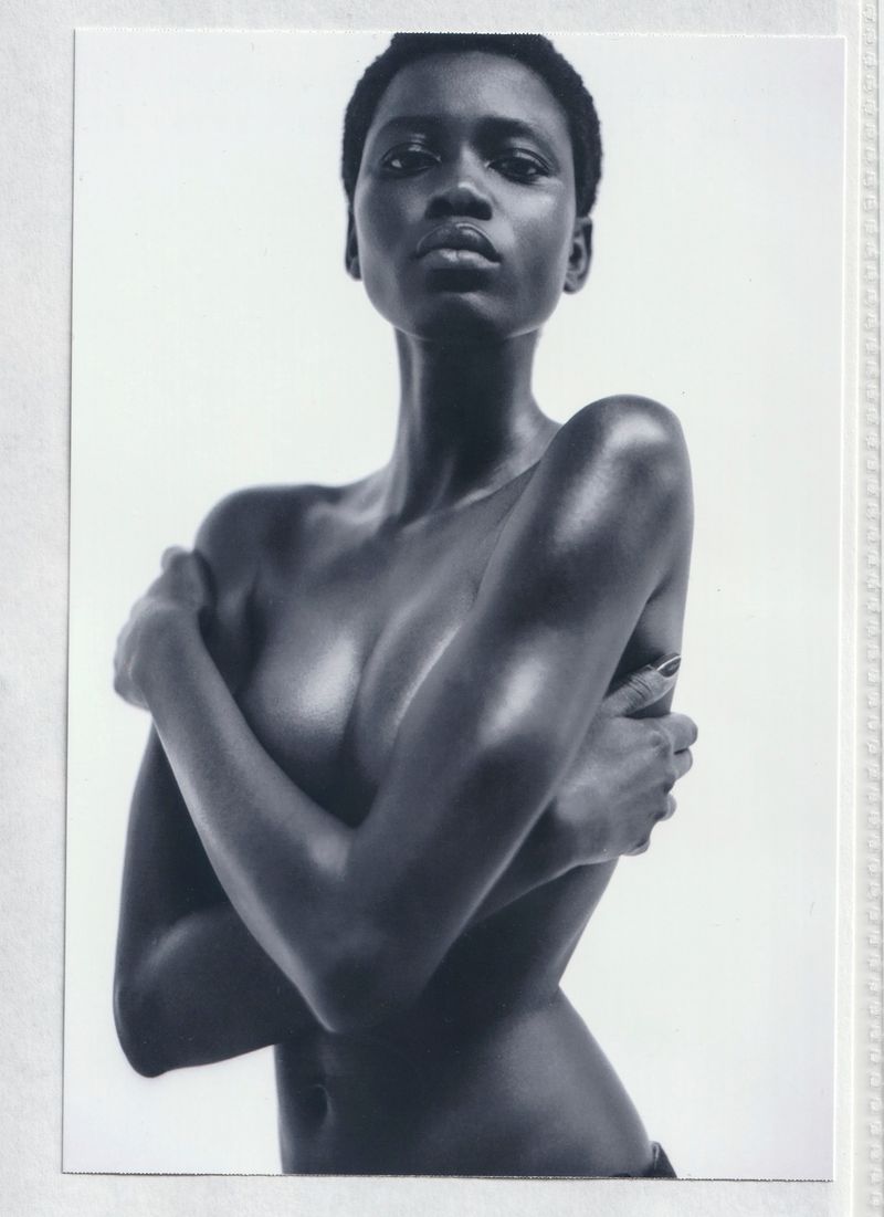 Black and white portrait of model Ajah Angau Jok posing topless with arms crossed against a white studio background.
