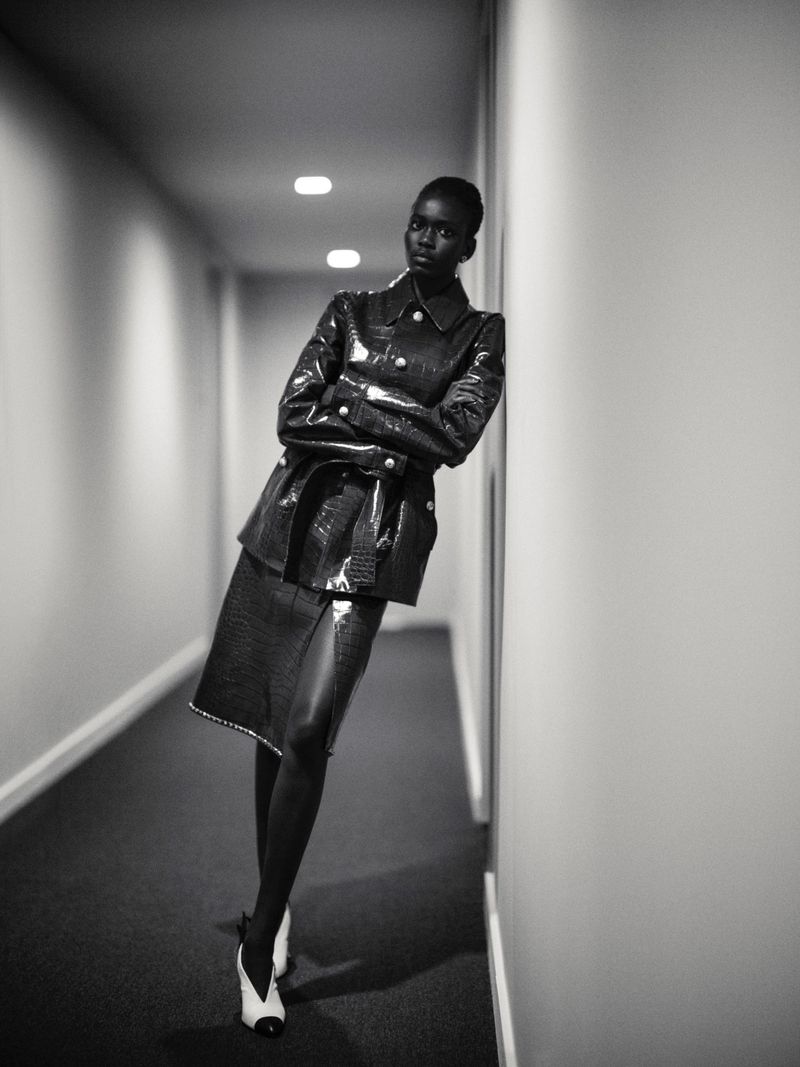 Model Ajah Angau Jok in a belted black leather jacket and skirt leaning against a hallway wall in black and white.