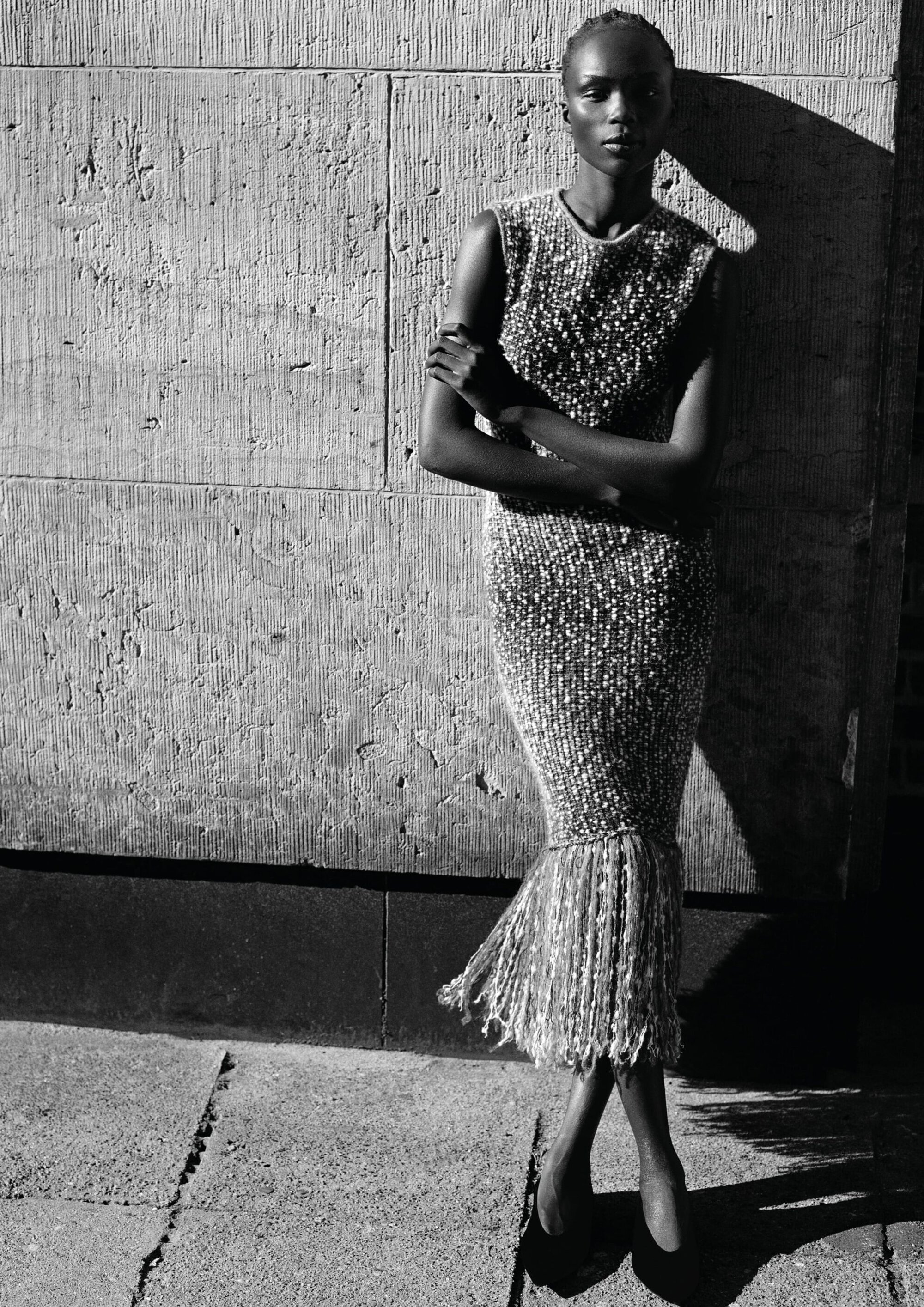 Model wearing a sleeveless embellished fringe dress posing against a textured stone wall in high-contrast sunlight.