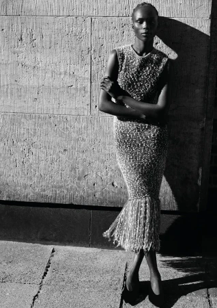 Model wearing a sleeveless embellished fringe dress posing against a textured stone wall in high-contrast sunlight.