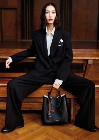 East Asian model in an oversized black suit sitting on a wooden bench holding a black leather tote bag on parquet flooring.