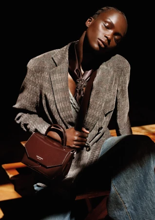 A Black woman in a textured grey blazer and wide-leg jeans holding a brown leather handbag against a dark background.