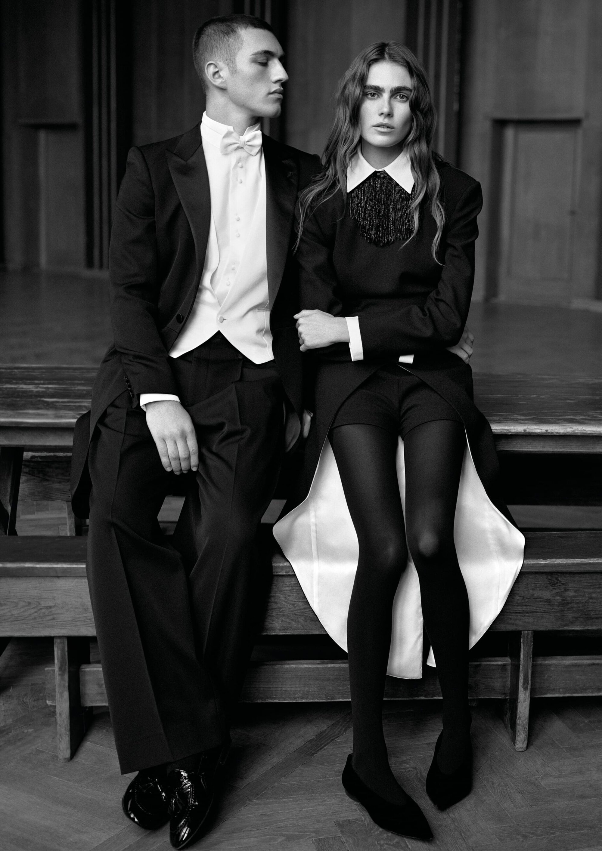 Man in a tuxedo and woman in a tailored black dress and tights seated together on a wooden bench in black and white.