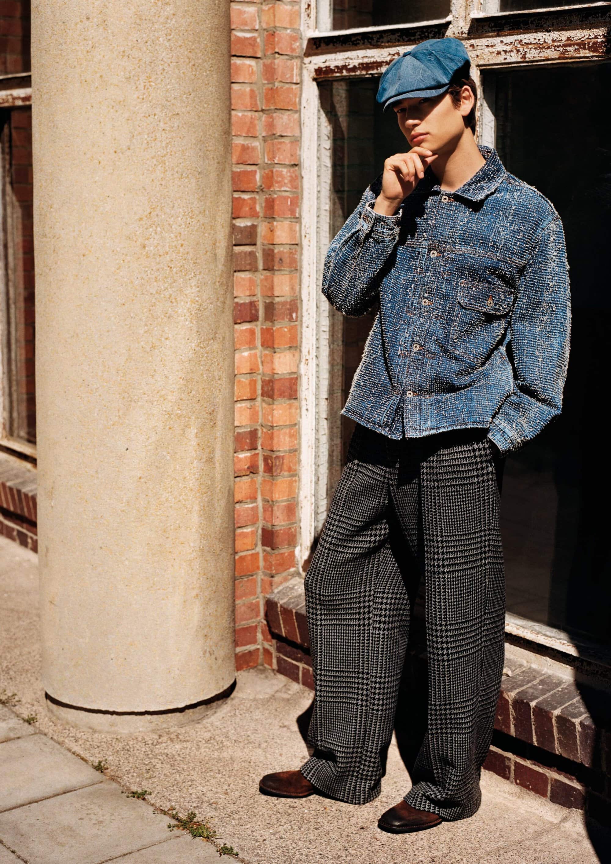 Model posing in a blue textured jacket, wide-leg plaid trousers, and a blue cap outdoors against a brick wall.