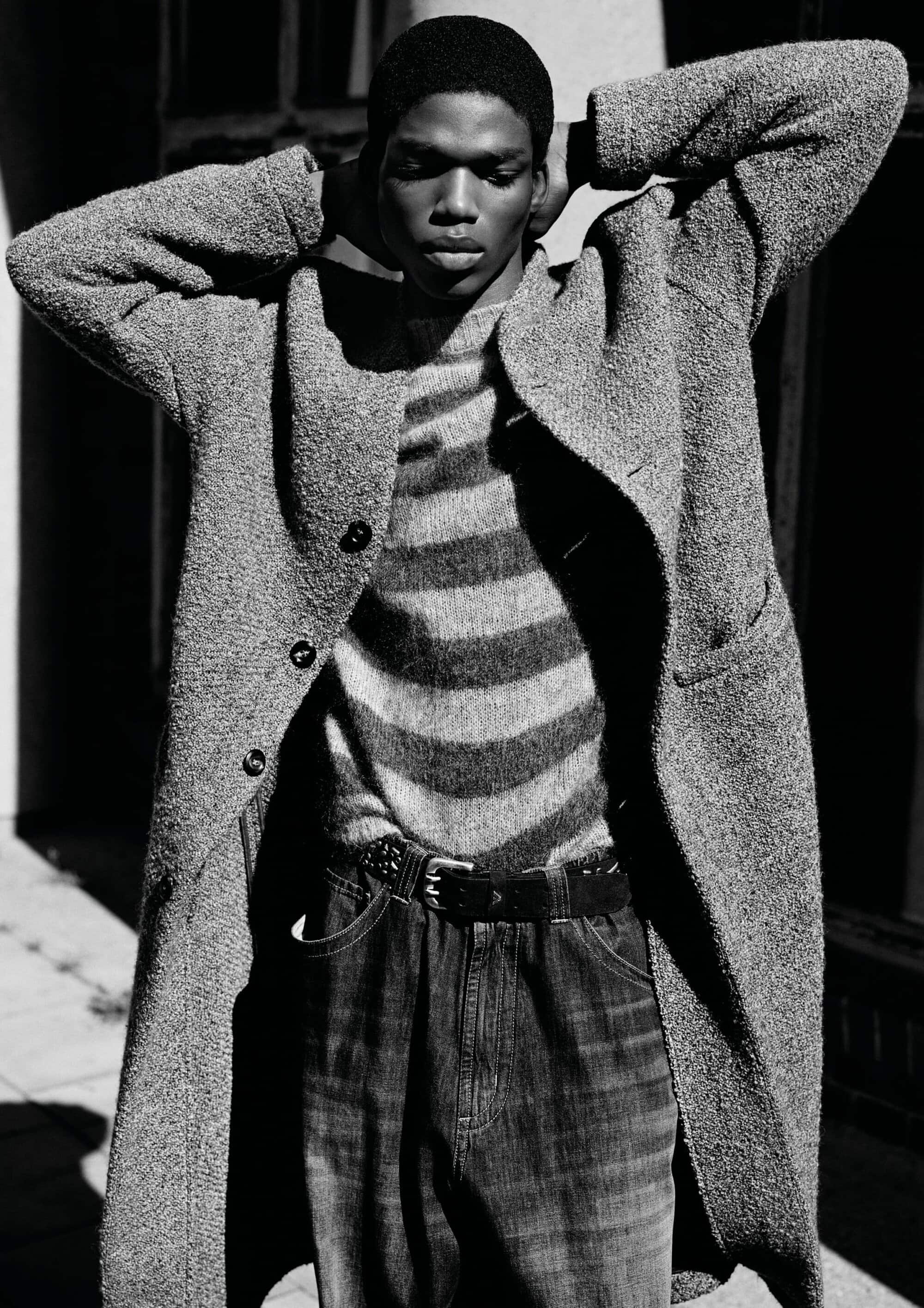 Black and white portrait of a Black male model wearing a textured overcoat, striped knit sweater, and belted denim trousers.
