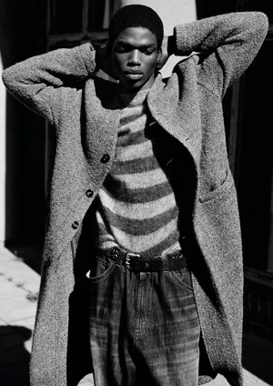 Black and white portrait of a Black male model wearing a textured overcoat, striped knit sweater, and belted denim trousers.