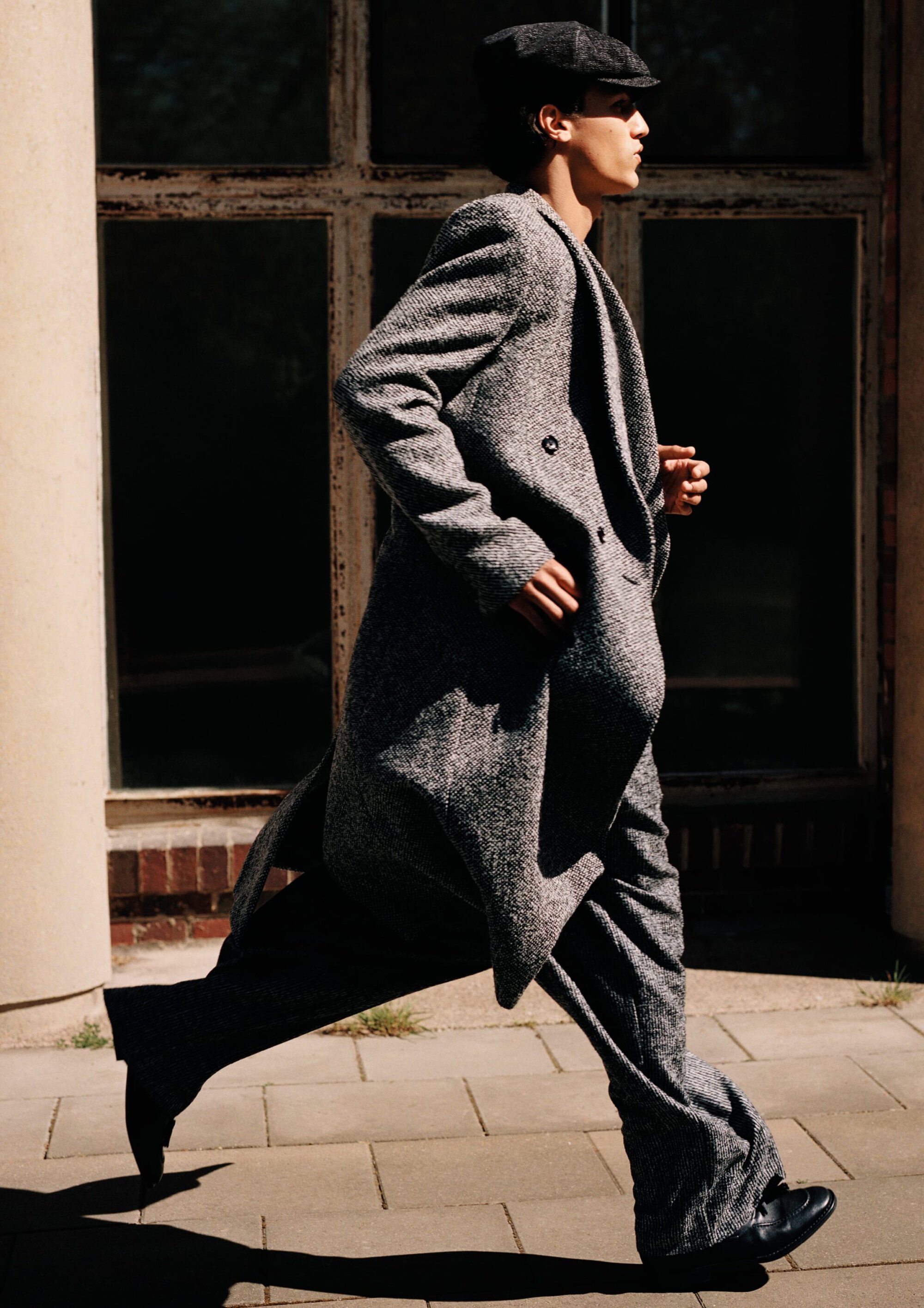 Young man in a grey tweed overcoat and flat cap running across a paved sidewalk in front of a building facade.