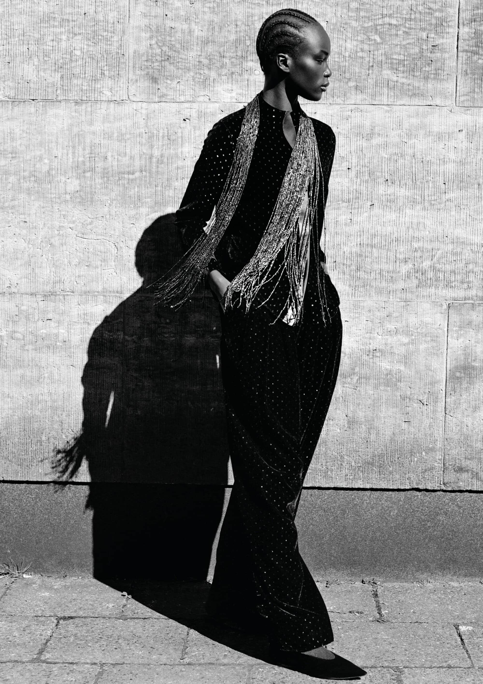 Black and white portrait of a model with braided hair in an embellished dark outfit and fringe scarf against a stone wall.