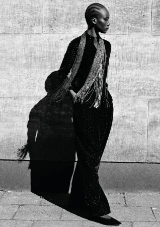 Black and white portrait of a model with braided hair in an embellished dark outfit and fringe scarf against a stone wall.