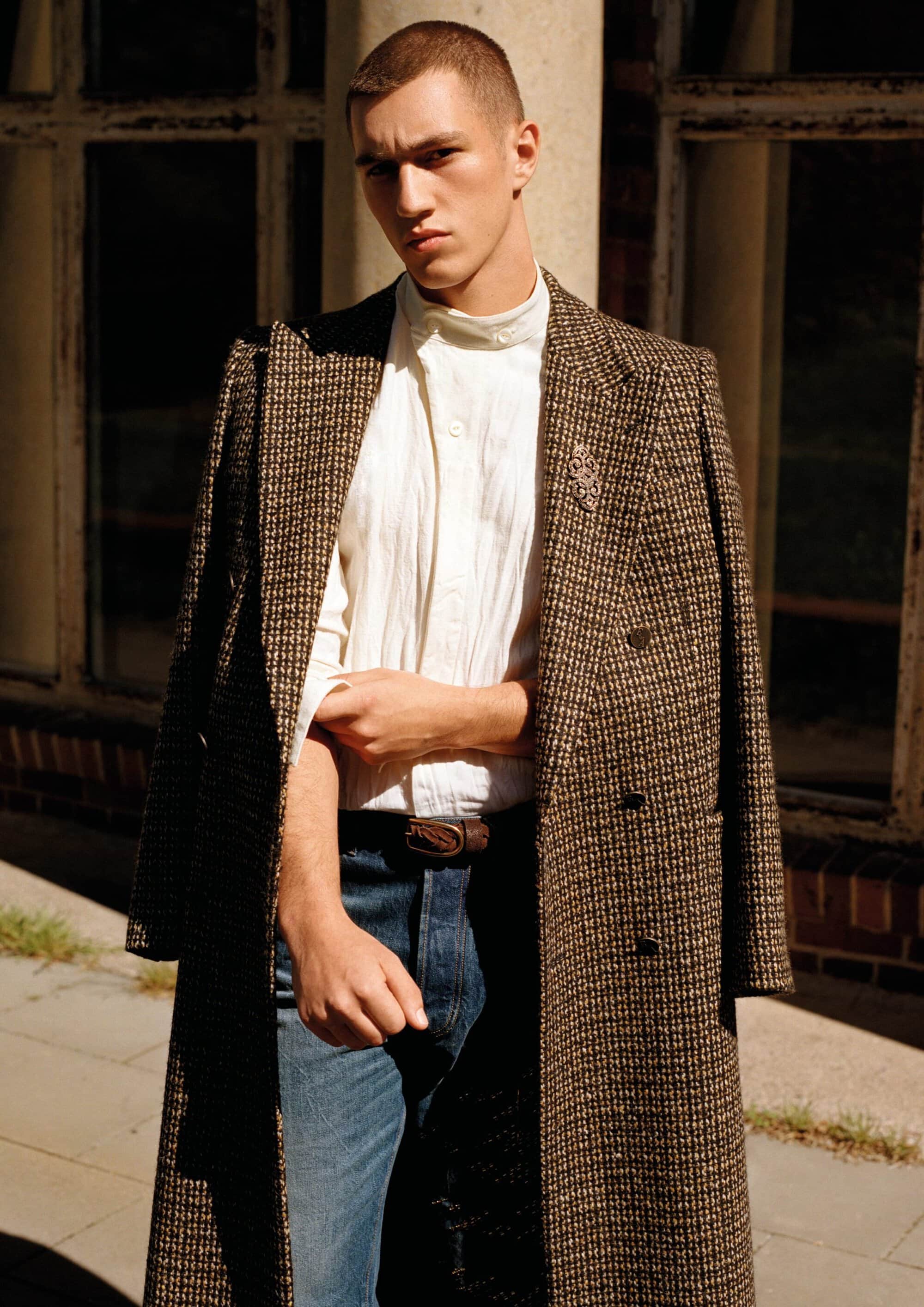Male model in a textured brown tweed overcoat, white high-neck shirt, and blue denim jeans standing in bright sunlight.