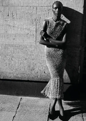 Black and white portrait of a woman standing in sunlight wearing a sleeveless sequined fringe dress against a stone wall.