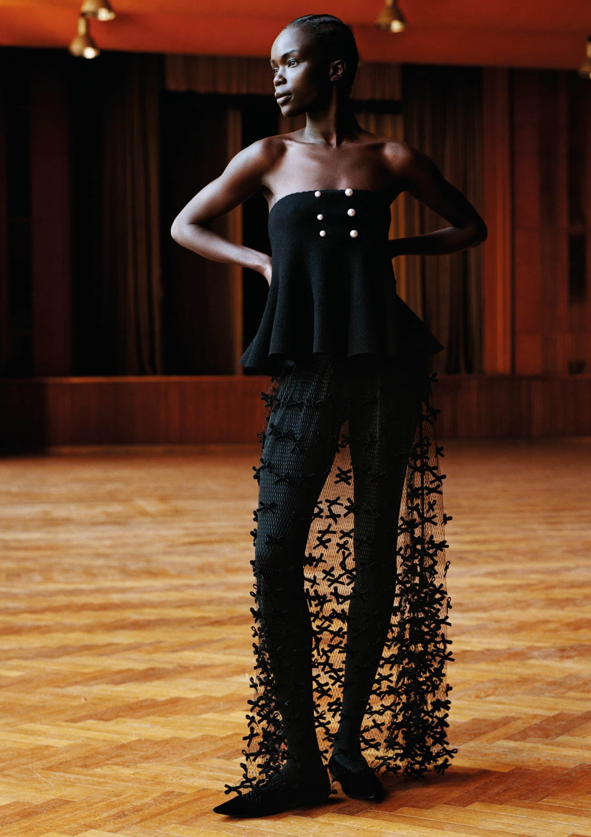 Model Jesephen Akuei posing with hands on hips wearing a black strapless peplum top and textured pants on a parquet floor.