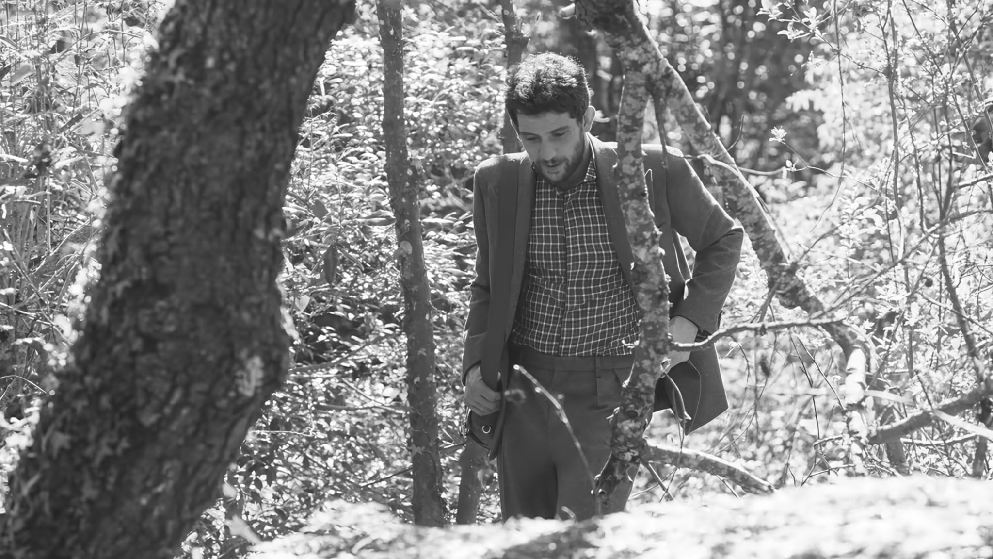Actor Josh O'Connor walks through dense woodland wearing a tailored suit and checked shirt in black and white.