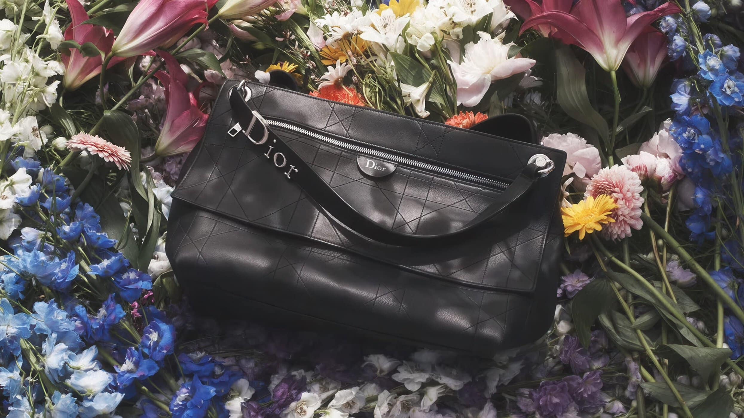 Black leather Dior handbag with Cannage geometric stitching resting atop an arrangement of vibrant colorful flowers.