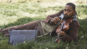 Lakeith Stanfield reclining on a grass field holding a violin beside a grey Dior messenger bag wearing a brown jacket.