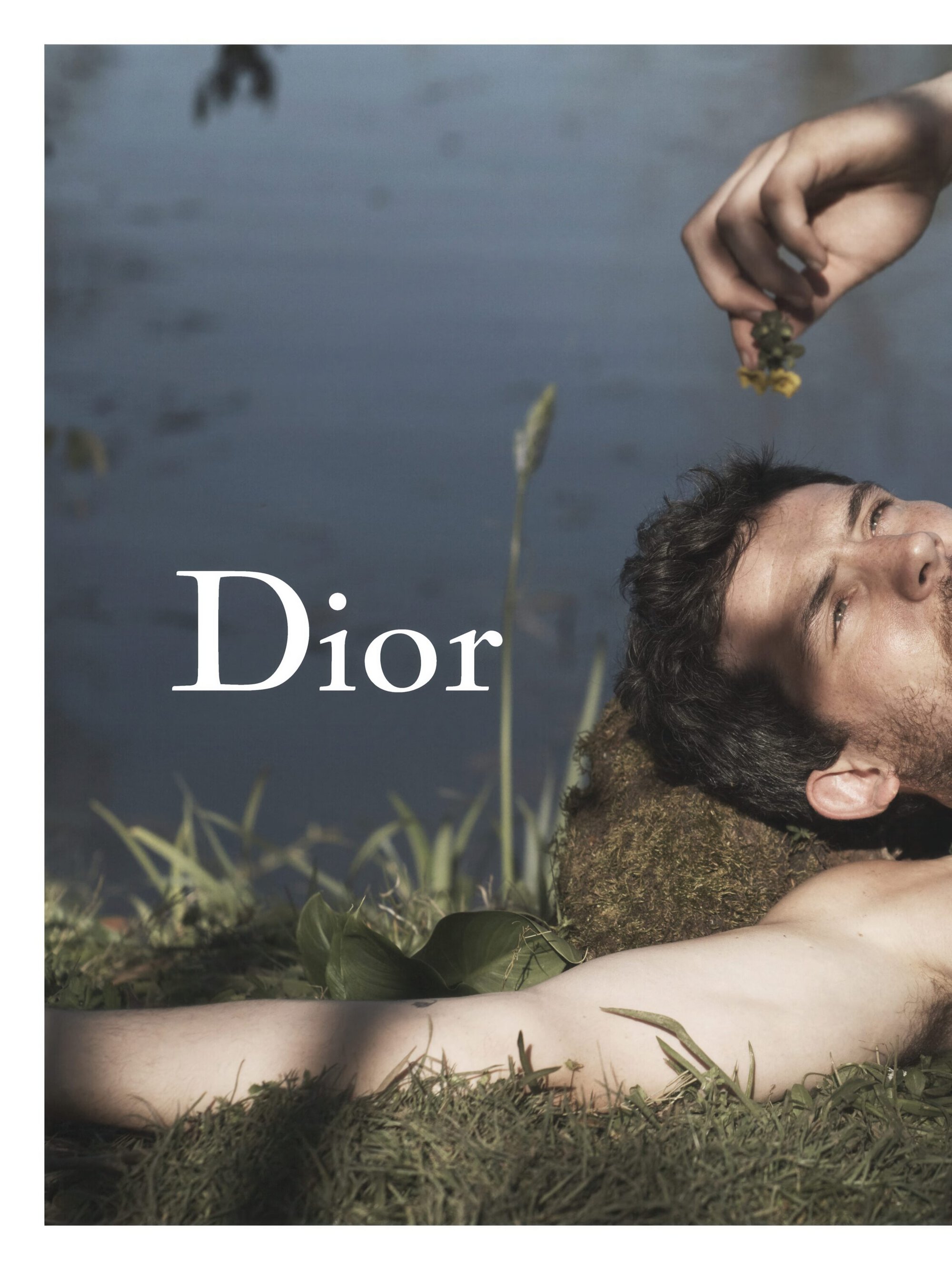 Josh O'Connor lying shirtless on grass beside water with a hand dangling a flower above him for Dior.