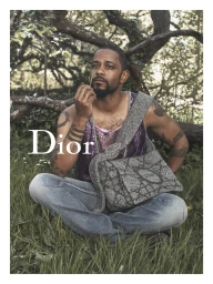 Lakeith Stanfield sitting cross-legged on grass wearing blue jeans, a purple tank top, and a grey textured Dior messenger bag.